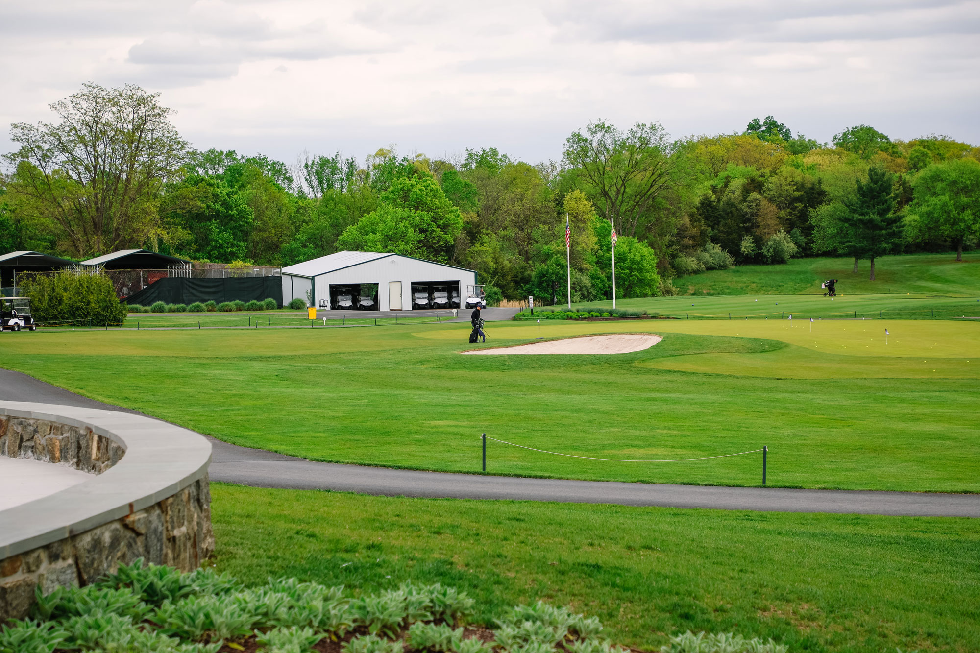 A person walks on a well-maintained golf course near a sand bunker, with a white building, trees, and greenery in the background under a cloudy sky.