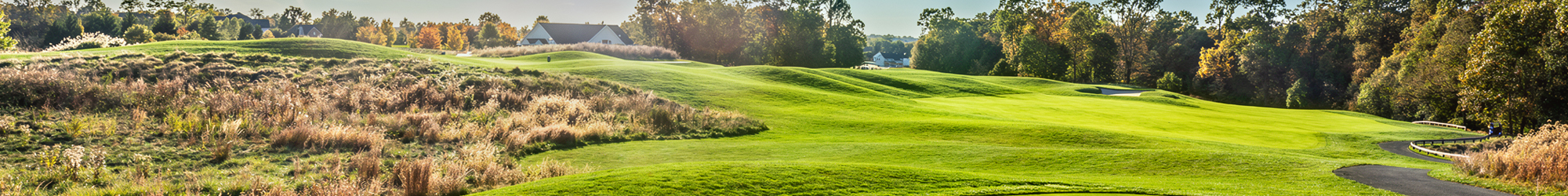 Lush green hills of Falls Road Golf Course with trees and a house in the background under a clear sky.