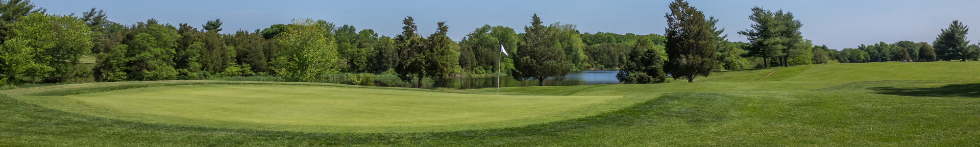 A panoramic view of a golf course with lush green grass, trees, and a pond in the background.