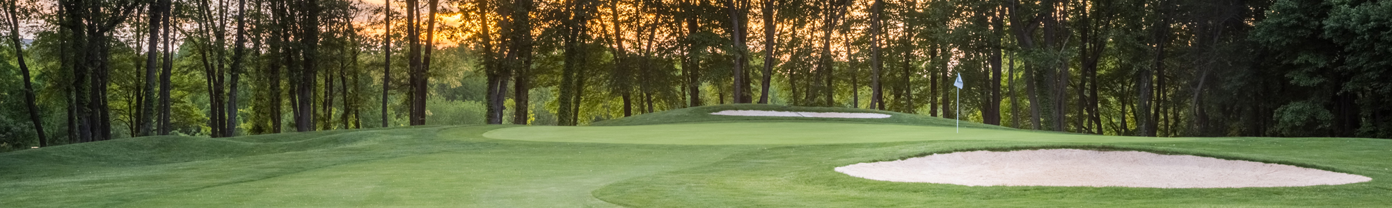 A scenic view of a golf course at sunset, featuring green hills and sand traps surrounded by trees.