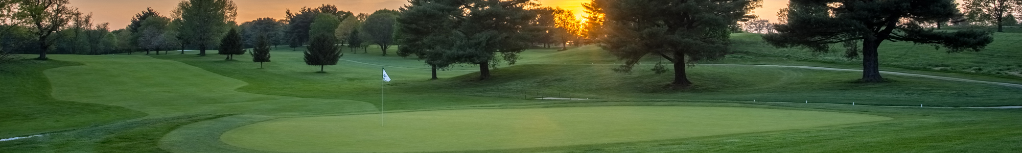 Sunset over a golf course with a green and flag, surrounded by trees and rolling hills.